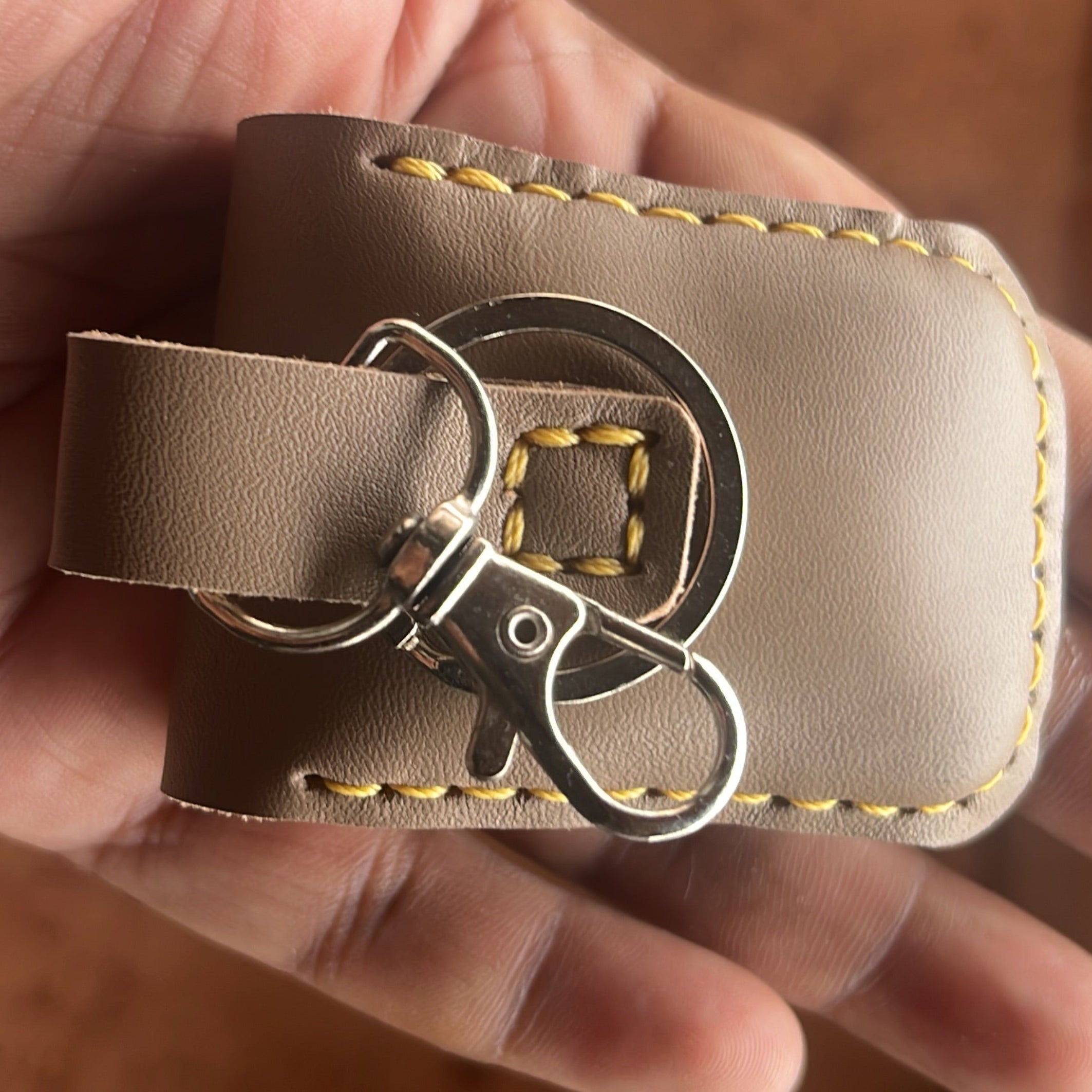 Hand holding a beige leather key case with a metal '&amp;' symbol on a wooden surface.